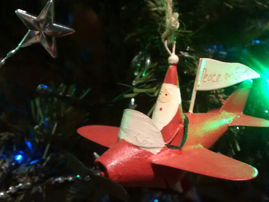 Close view of decorations on an artificial Christmas Tree.  In the top left there is a silver star.  Most of the right side of the picture is taken up by a rather conical Father Christmas piloting a bright red monoplane. The plane flies a 'Peace on Earth' banner.