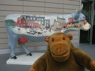 Mr Monkey in front of a cow decorated with a cow-filled Lowry-like painting
