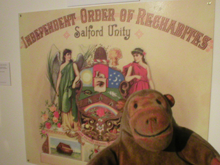 Mr Monkey looking at a poster showing the arms of the Independent Order of Rechabites Salford Unity