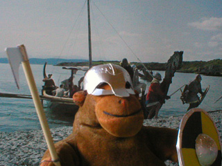 Monkey in his Viking helmet, with a warband and longship on the beach behind him
