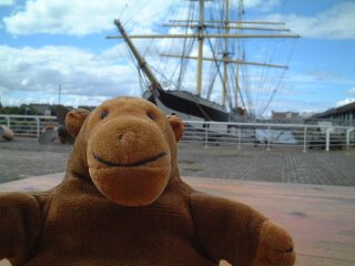 Mr Monkey on a dock, with a sailing ship behind him
