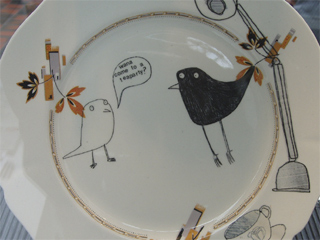 A small white bird invites a black bird to a tea party on a plate painted by Rachael Gardiner