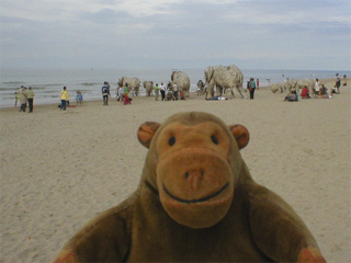 Mr Monkey looking at a group of wooden elephants surrounded by people